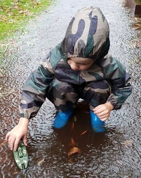 Small child wearing Mud Mates NZ-made waterproof camo coveralls playing in puddles with a toy boat