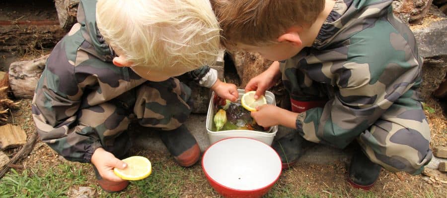 Nature Play Activities for Toddlers & Preschoolers: 2 young boys wearing Mud Mates camo coveralls placing natural treasures into a container to freeze for ice play